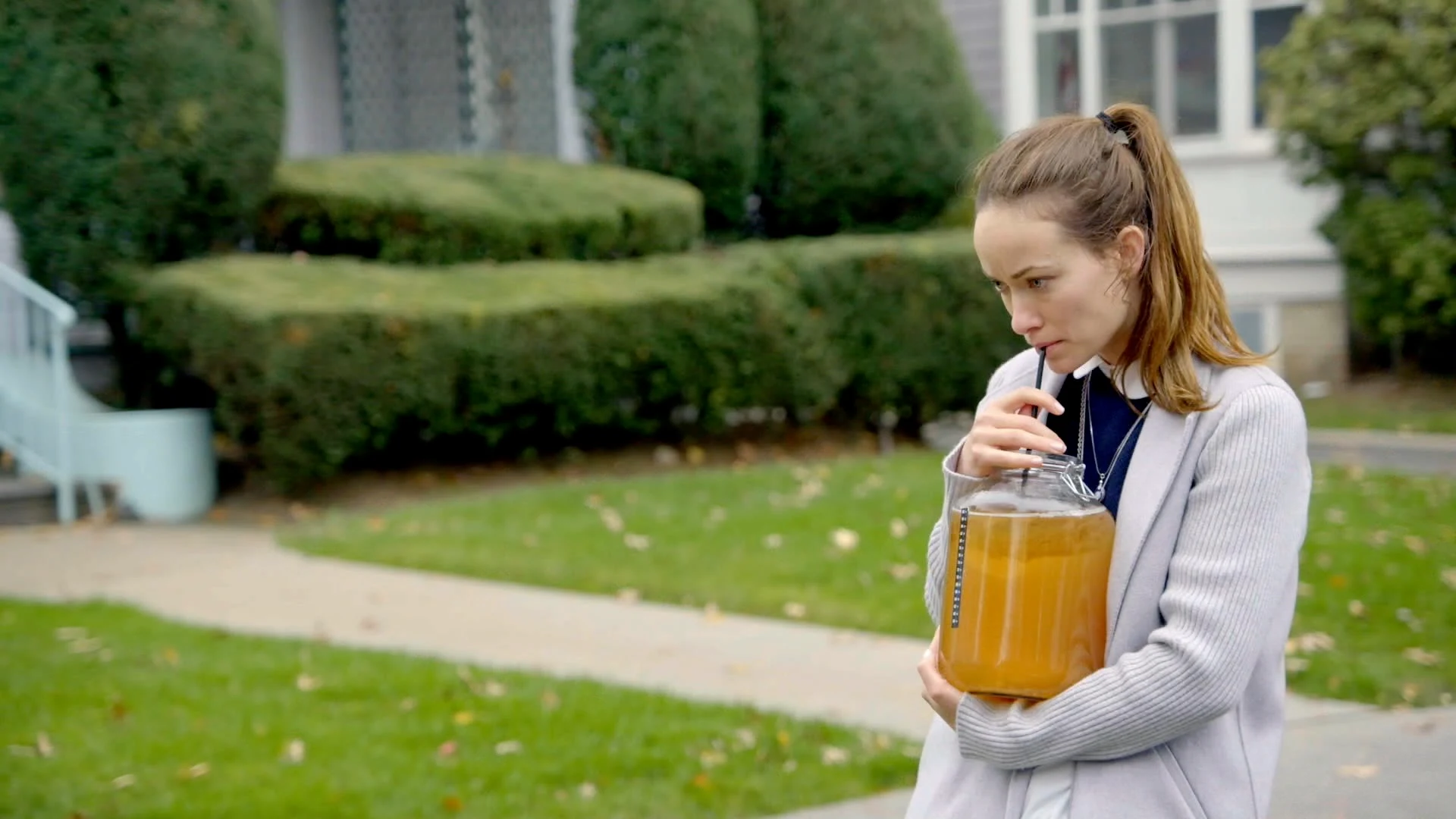 Olivia Wilde holding a large jar of kombucha outside a house in Daytime Noon.